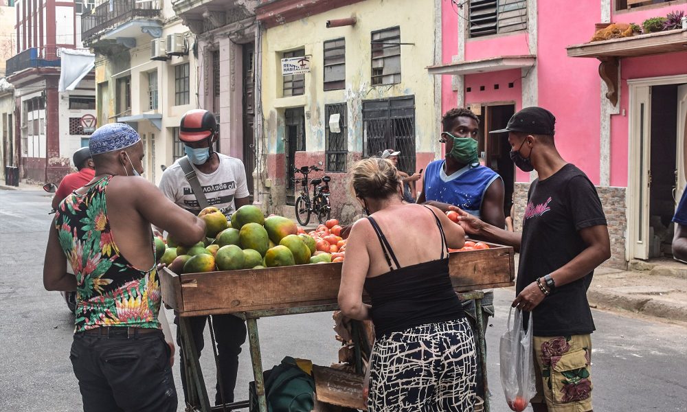 Varias personas compran frutas y vegetales en un puesto callejero en Cuba. La carretilla, cargada con mangos y tomates, refleja el acceso limitado y estacional a los alimentos en el país.