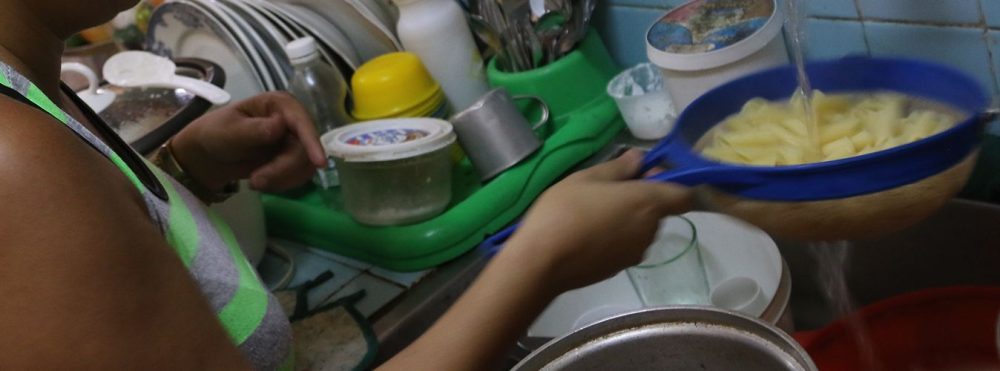 Una mujer escurre pastas en un colador azul sobre un fregadero lleno de utensilios y recipientes plásticos.