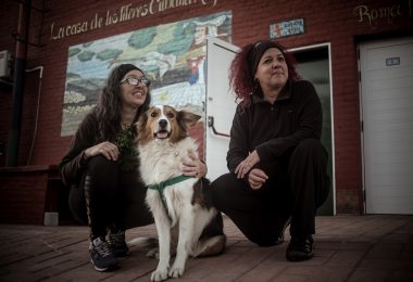 Geraidy Brito Montes de Oca y Yaqui Saiz, junto a su perro, sentadas frente al mural de la Casa de Títeres Cubana Argentina en Junín. Detrás, se ve la fachada del espacio cultural con puertas blancas y paredes de ladrillo rojo. Foto: Ella Fernández.